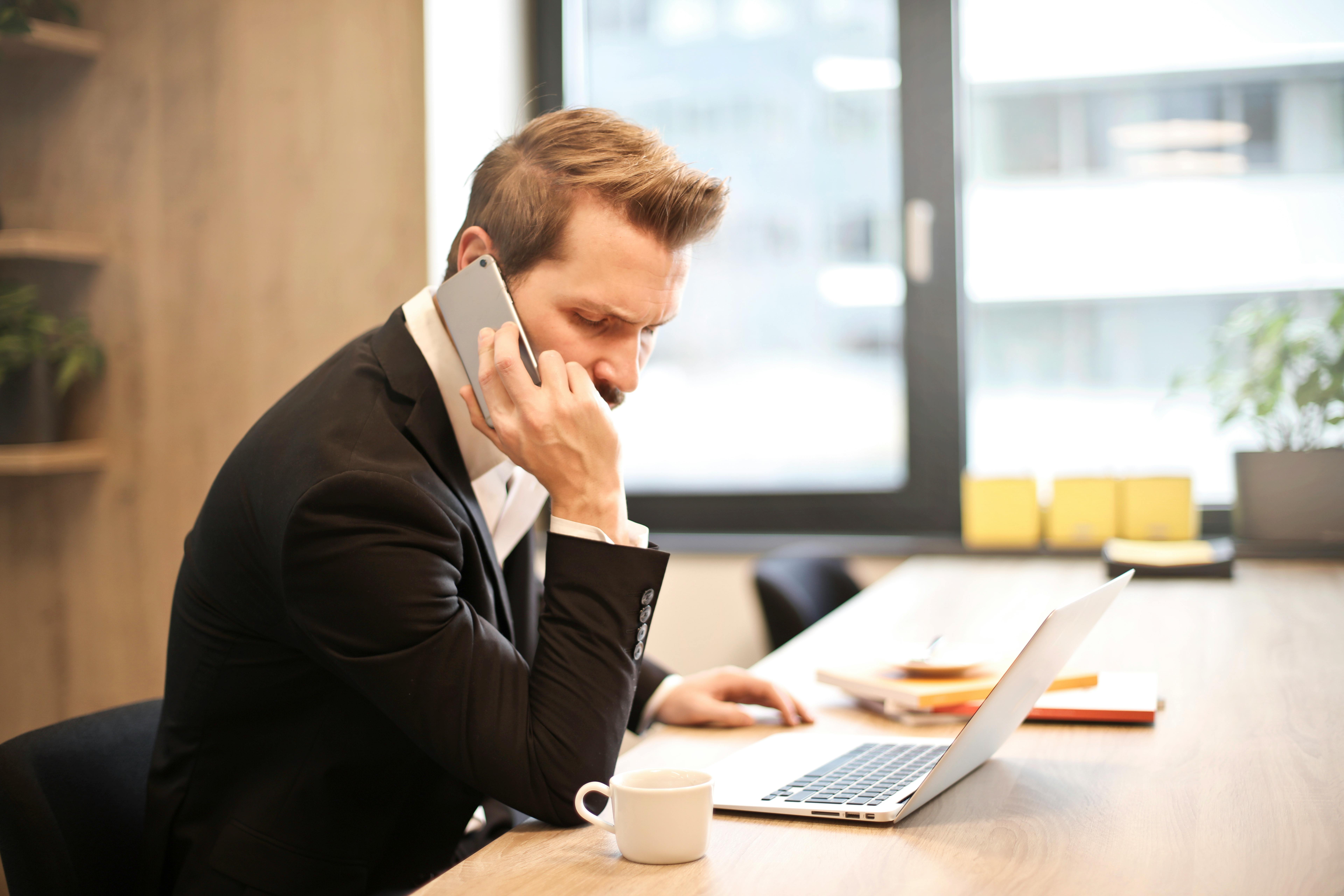 A man in a suit sitting at a table while talking on his cellphone.